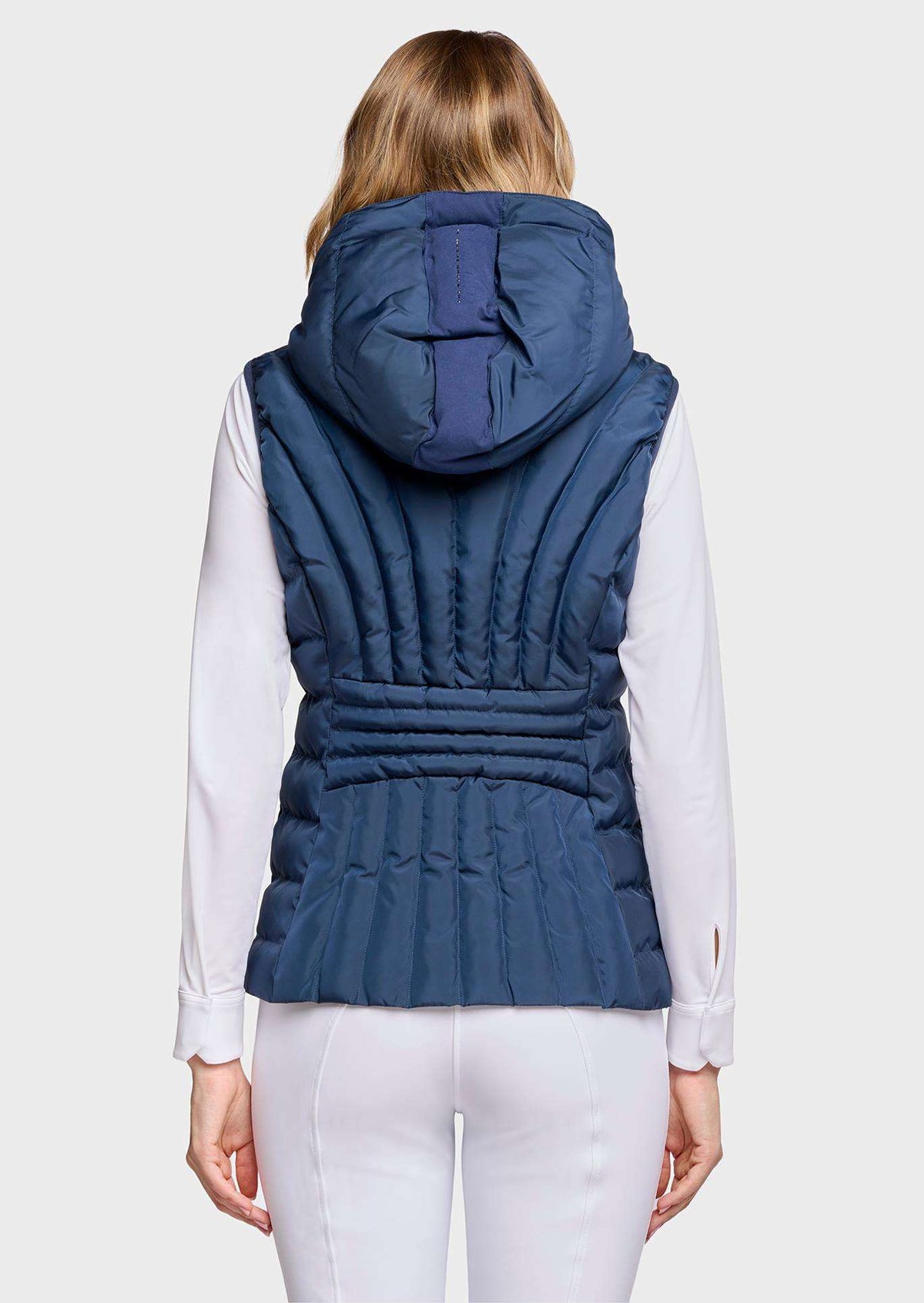 A woman viewed from the back wearing a navy blue hooded puffer vest over a white long-sleeve top and white pants, against a light gray studio background.