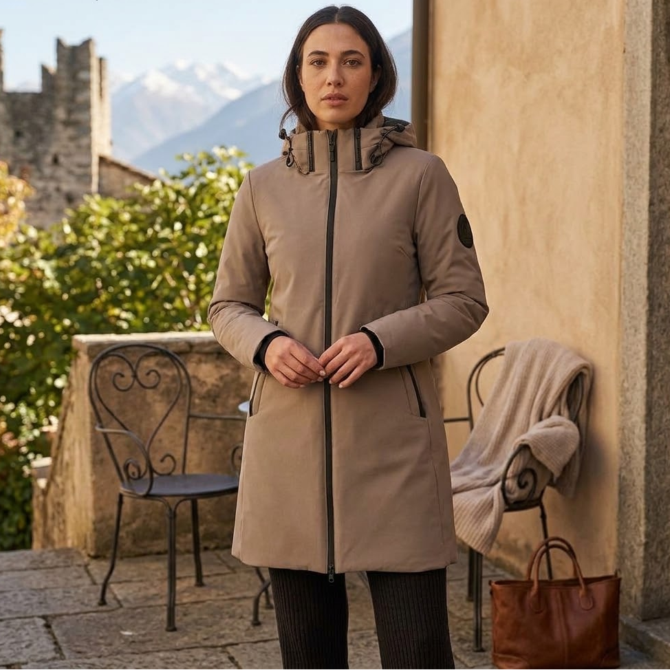 A woman stands outdoors wearing a mid-length beige hooded jacket with a full front zipper and zip hand pockets. The setting includes stone walls, chairs, a draped knit blanket, and mountains in the background.