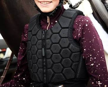Person wearing a black equestrian helmet and protective vest standing next to a horse.