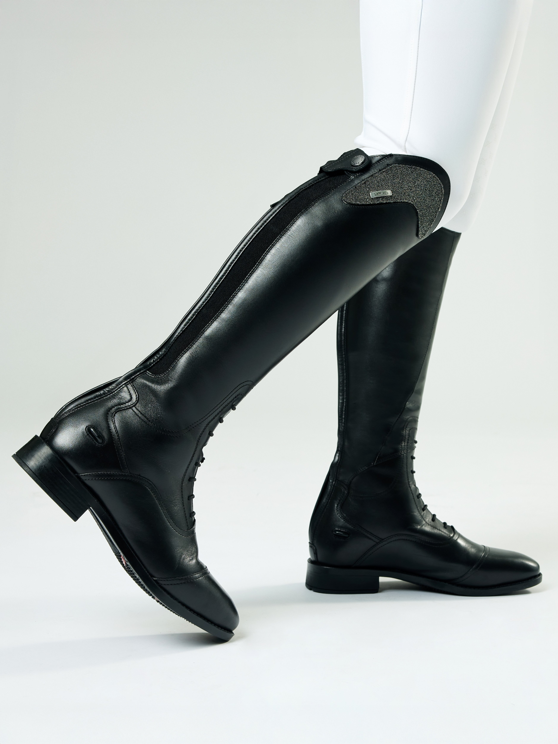 Black riding boots worn with white pants on a light gray background