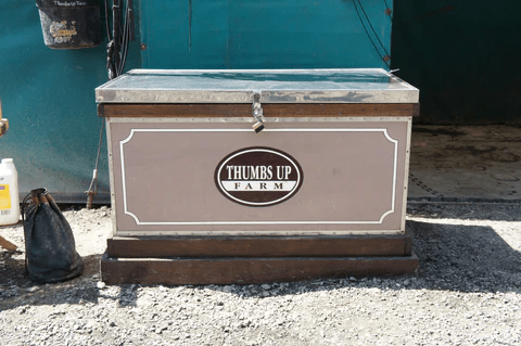 A wooden chest labeled 'Thumbs Up Farm' on gravel with a teal background.