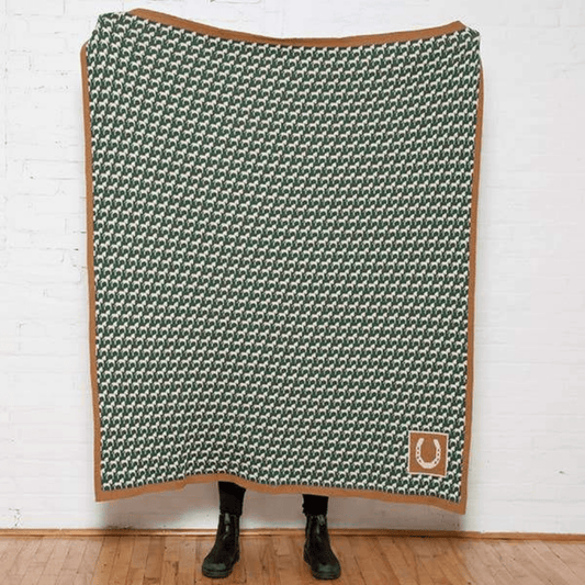 In2Green - Eco Lucky You Equestrian Throw held by a model against a white wall and wood floors. Hunter green horse-houndstooth, tan border, and tan horseshoe icon. 
