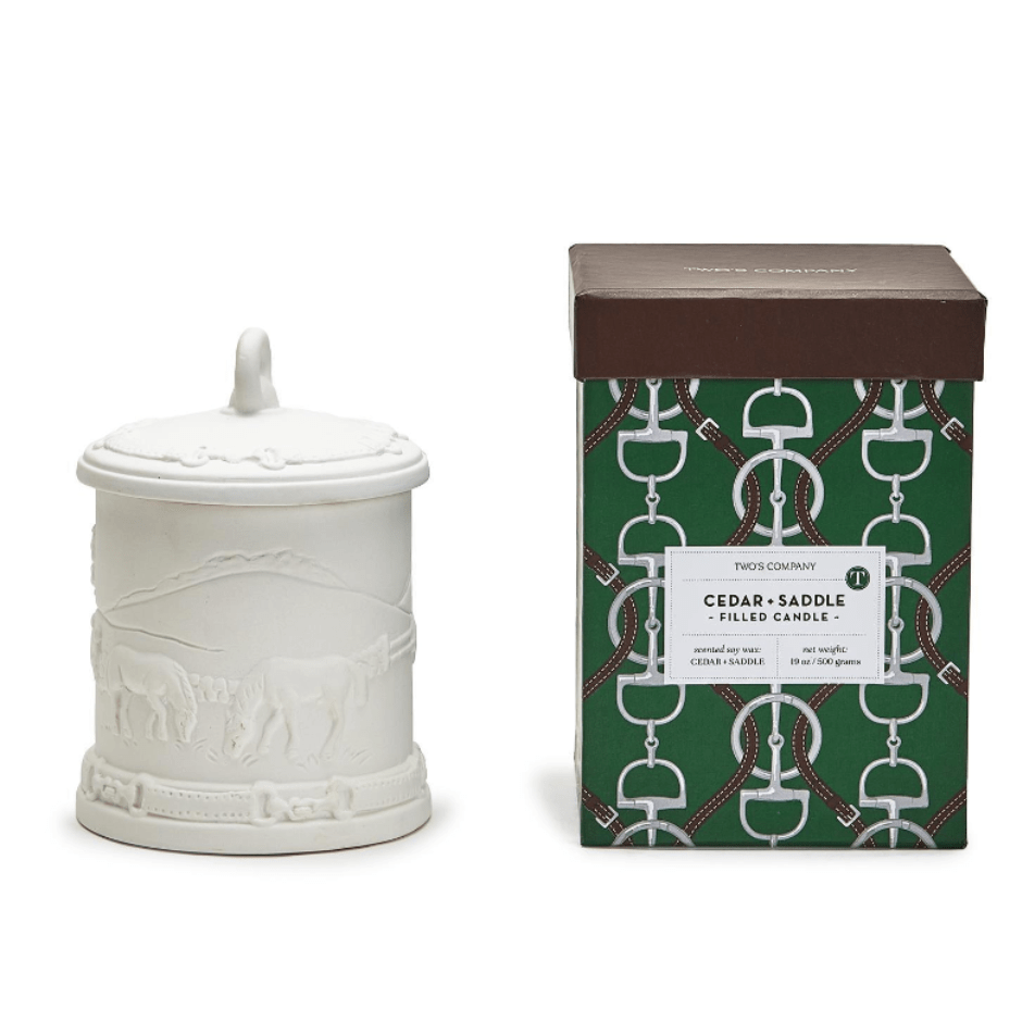 White ceramic candle votive decorated with horses and a lid, next to a decorated green & brown gift box labeled Cedar & Saddle Filled Candle