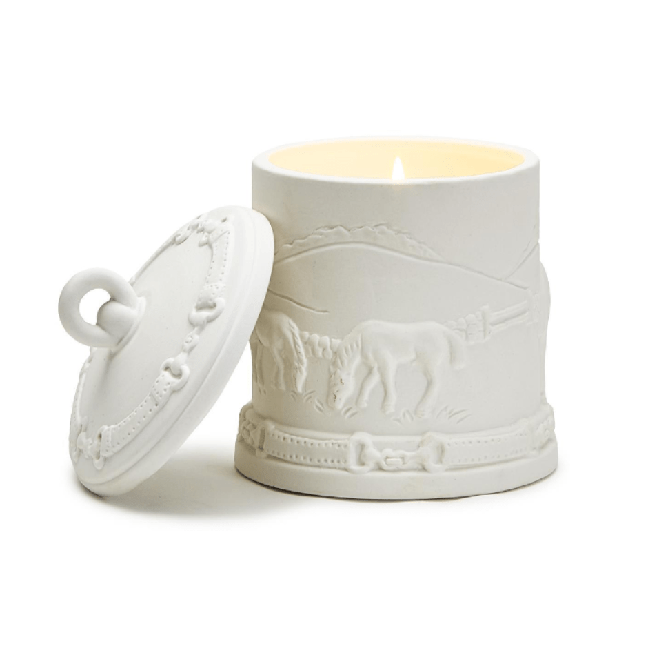 White ceramic candle votive decorated with 2 horses in a field. The lid leans against the left side. 