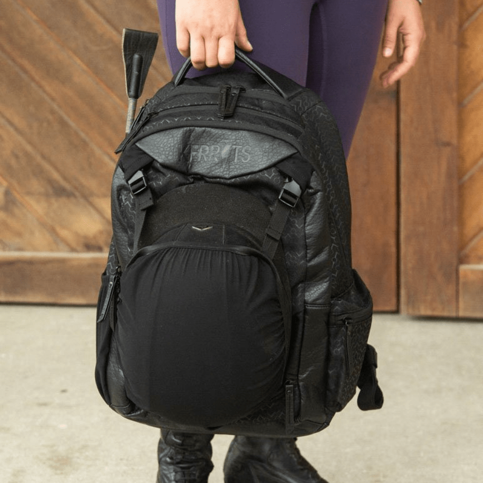 Model holding a black Kerrits backpack in their right hand at thigh height. The backpack is zipped up with a whip in the right pocket and a helmet in the front mesh pocket. Wood wall and concrete floor in the background. 